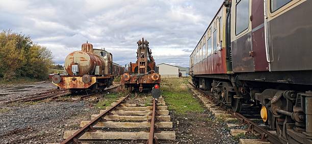 Old fashioned locomotive engines, wrecked train carriages, rusting railroad tracks. Rusted railway remains, derelict vintage steam engine and railcars. Post-apocalyptic scene of decaying infrastructure. Abandoned rail siding transport yard with boxcar ruins and corroded passenger car carriage. Trainyard storage for obsolete weathered boxcars, cargo, engine, and freight trains to decay. Railcar relics of the past graveyard.