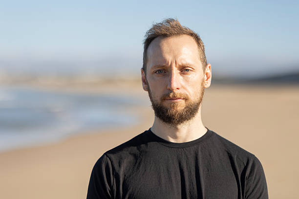 Adult man with a beard standing on a sandy beach looking straight ahead