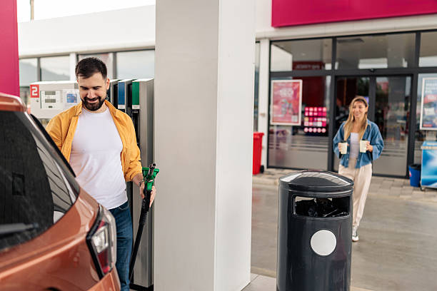 Woman carrying two takeaway coffees from the gas station store as her boyfriend fills the car with fuel.