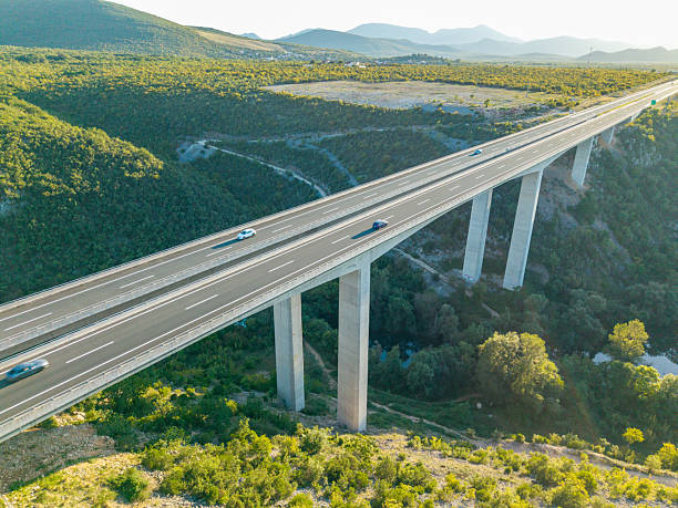 Aerial view of a modern elevated highway bridge spanning a lush green valley with light traffic. Scenic landscape with mountains in the distance and a clear sky. Infrastructure and nature.