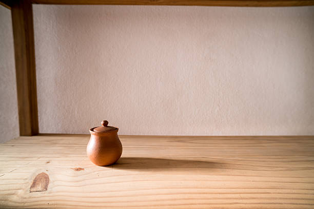 A simple, lidded pot on a wooden shelf, against a soft, textured wall background.