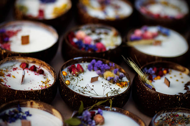 Hand-poured candles decorated with dried flowers and herbs in coconut shell containers arranged on a table.