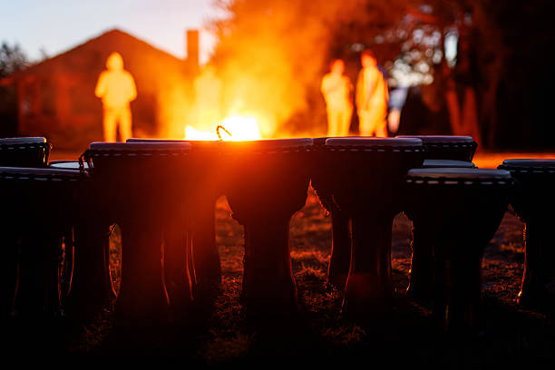 Silhouetted drums in the foreground with people standing near a bright campfire in the evening.