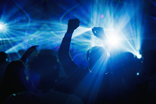 Group of silhouettes raising hands and dancing under strong blue laser beams during a live music concert.