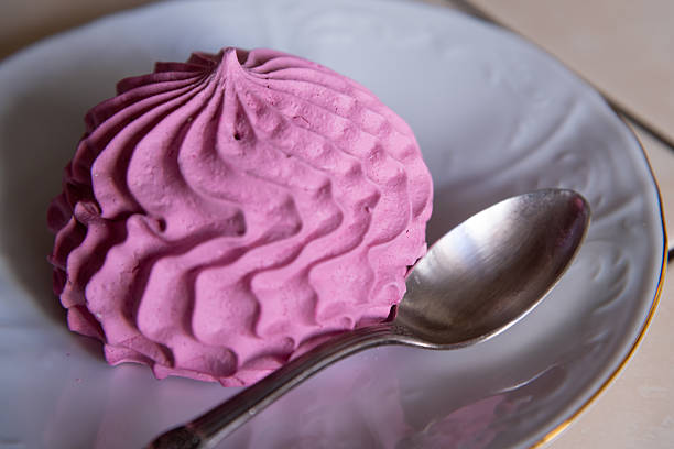 A fluffy pink dessert sits on a white plate, featuring a textured surface. A shiny silver spoon rests beside it, inviting a taste. This delightful treat looks both appetizing and elegant.
