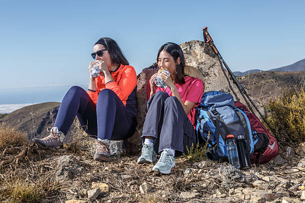 Two women sit against a rock on a mountain trail, enjoying snacks and hydration after hiking. The clear blue sky and scenic landscape reflect a sunny outdoor adventure in nature.