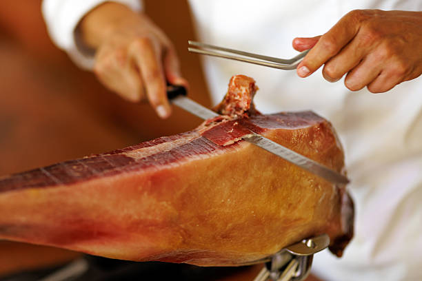 Close-up of a chef slicing cured ham leg with a long knife and precision in a kitchen setting.