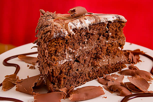 Close-up, wide slice of chocolate cake, two layers in the middle with chocolate cream, topped with meringue glaze lightly mixed with chocolate, surrounded by light chocolate shavings and a drizzle of chocolate decorating the white plate, red background.