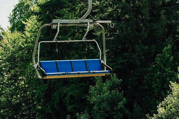 An empty ski lift chair swings gently in the air set against a backdrop of vibrant green trees under a clear blue sky. It captures the peacefulness of a summer day in the mountains.