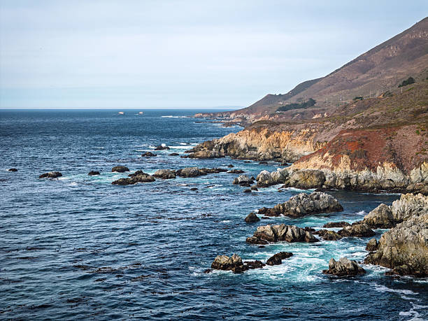 Aerial view of the Pacific Ocean coastline south of Carmel-by-the-Sea, near Garrapata Beach in Big Sur, California. Rugged cliffs and ocean waves under a bright, clear sky.