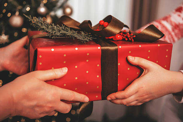 istock Two hands are passing a red and gold gift box adorned with a bow and greenery, cozy setting includes a decorated Christmas tree in the background 2244246136