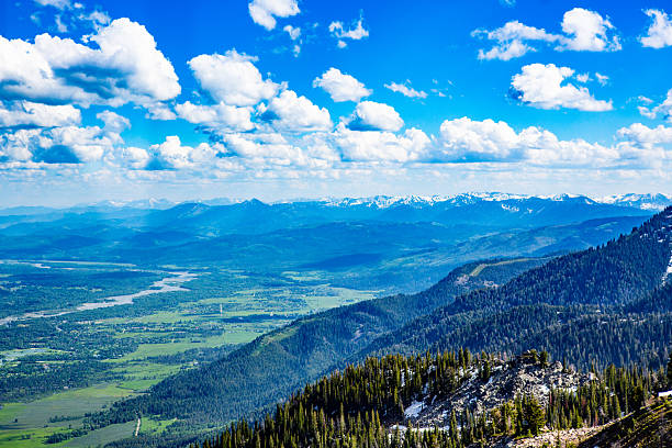 From the heights of the Teton Range, the world opens below — a sweeping valley of rivers, forests, and farmland unfolding toward a blue horizon. The Snake River glistens like a silver thread, winding through the lush basin far below. Snow-capped peaks frame the distant skyline beneath an ocean of sculpted clouds and cobalt sky. This panoramic image captures the full majesty of Wyoming’s alpine perspective — where altitude meets atmosphere and every mile of wilderness feels infinite.