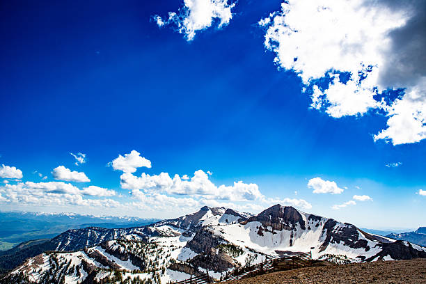 A breathtaking alpine vista captures the Teton Range stretching toward an endless horizon. The snow-streaked ridges rise from emerald foothills into a sky of pure sapphire blue, dotted with drifting white clouds. Shafts of sunlight break through the upper clouds, creating visible rays that fan across the scene like divine brushstrokes. The weather-worn wooden snow fence traces the ridge line below, anchoring the composition against the vast celestial expanse. This image embodies the clarity, silence, and magnitude of high-mountain air — a visual meditation on altitude and freedom.