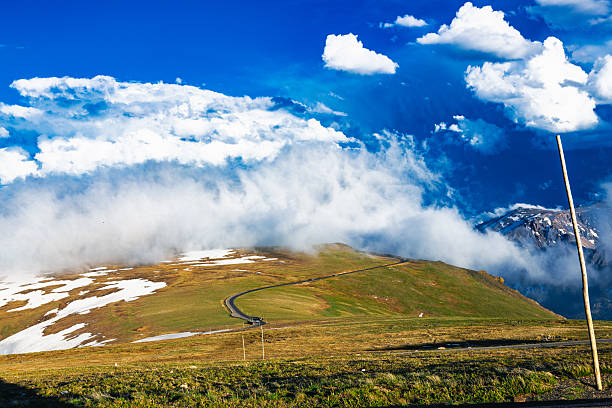 A serpentine alpine road winds through patches of melting snow and open tundra, disappearing into a cascade of rolling clouds. The earth below glows with golden light while the heavens above churn in luminous blue and white. The horizon dissolves into mist, erasing the line between mountain and sky. This image captures a rare moment on the Continental Divide where the world feels suspended — neither earthbound nor celestial, but beautifully caught in between.