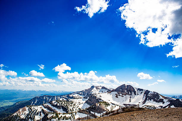 An elevated view over the Grand Teton Range reveals an endless horizon of snow-covered ridges and distant blue valleys. A vast sky, rich with cobalt gradients and soft clouds, stretches infinitely above, framing the high mountain world below. Sunlight scatters through the clouds, casting beams of light across the rugged slopes and deep snowfields. A weathered snow fence lines the foreground, symbolizing both isolation and endurance at the roof of Wyoming’s wilderness. The image captures the feeling of standing at heaven’s edge — where land, light, and air converge in absolute stillness.