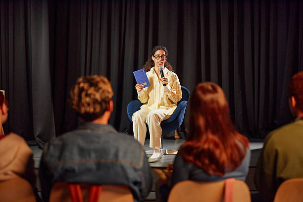 istock Young Adult Woman Hosting Public Reading Event on Stage Facing Audience 2244254601