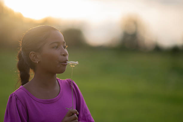 A smiling young Black girl in a purple shirt blows on a dandelion in a sunlit outdoor field, capturing playful innocence and warm, golden-hour glow. A natural, carefree childhood moment.