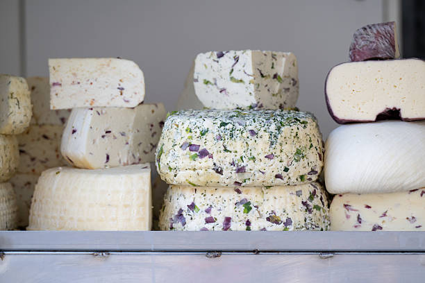 A colorful display of assorted artisanal cheeses stacked on a counter, featuring a variety of textures, shapes, and added herbs or spices. Some cheeses are flecked with purple onion and green herbs, while others show smooth or aged rinds. The image reflects traditional cheese-making and gourmet food culture.