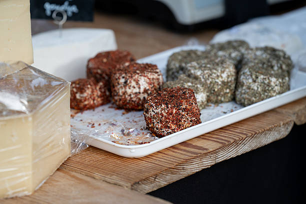 Cubes of artisanal goat cheese coated in various spices, including red pepper and dried herbs, are displayed on a white tray at a market stall. The rustic wooden table and natural lighting highlight the textured surfaces and rich colors, evoking a gourmet and handcrafted food experience.