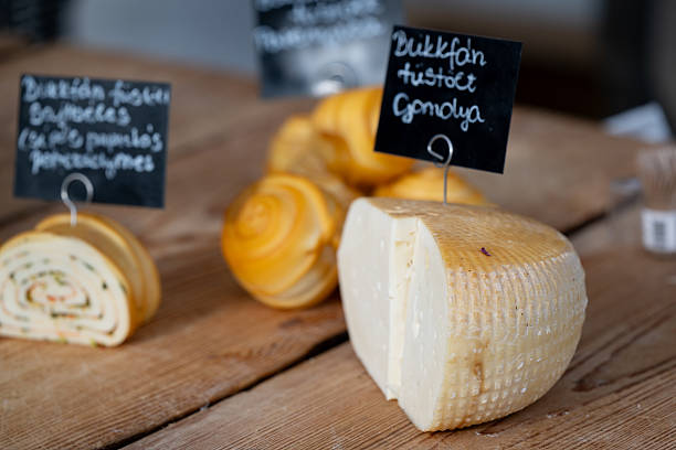 Sliced rolls of smoked cheese filled with herbs, onions, and spices are neatly arranged on a wooden counter at a market stall. Each variety is labeled with handwritten signs, highlighting the artisanal and local nature of the products. The spiral patterns and rich colors emphasize traditional cheese-making techniques.