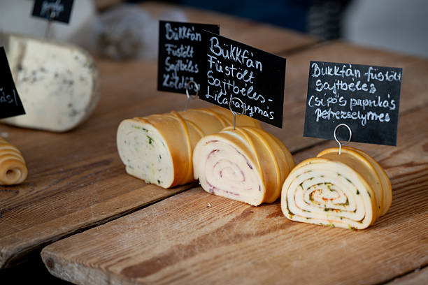 Sliced rolls of smoked cheese filled with herbs, onions, and spices are neatly arranged on a wooden counter at a market stall. Each variety is labeled with handwritten signs, highlighting the artisanal and local nature of the products. The spiral patterns and rich colors emphasize traditional cheese-making techniques.