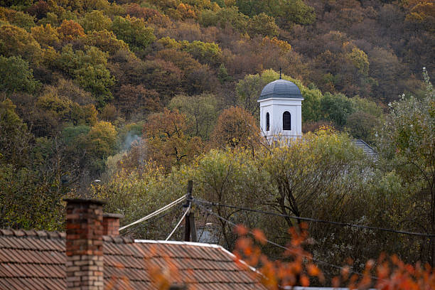 A white church tower with a dark dome rises above the treetops in a hillside village, framed by dense autumn foliage in shades of green, yellow, and brown. Rooftops and power lines in the foreground add a rustic, inhabited feel to the otherwise serene and natural setting.