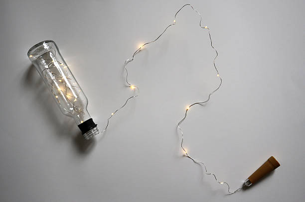 Cork shaped LED string lights spilling out of a clear glass bottle on a neutral gray background. Themes of home decor.