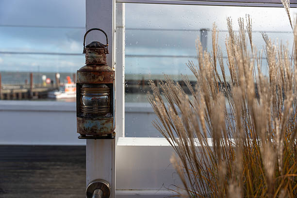 A rusty nautical lantern hangs beside tall grass near the harbor. The background shows glass panels with raindrops and a blurred view of the sea.