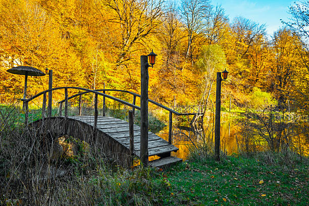 A wooden bridge over a river with a lamp post on each side. The bridge is surrounded by trees and the water is calm. The scene is peaceful and serene