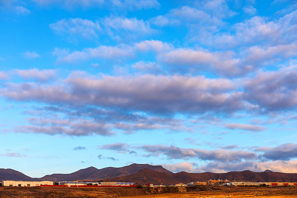 Landscape with cloudy sky during sunrise, range of mountains and some buildings visible in foreground. Similar landscapes are commonly found in locations like Canary Islands and Arizona and New Mexico