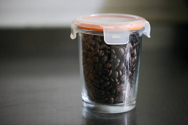 A transparent glass container with an orange lid filled with roasted coffee beans, suitable for storing coffee.