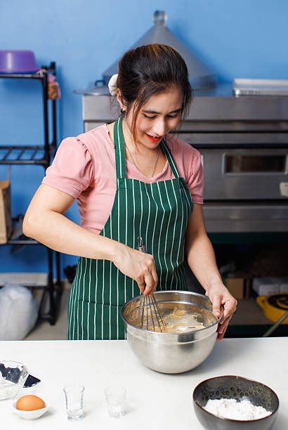 Asian beautiful young woman bakers wearing striped aprons whipping mixing flour different ingredients in metal bowl making dough baking process homemade bread pastry recipe in bakery kitchen concept