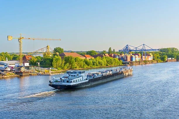 Mainz, Germany - June 19, 2025: Cargo vessel navigating the river in Mainz, Germany, with colorful shipping containers and cranes visible along the harbor under clear blue skies.