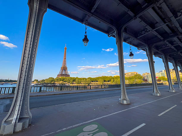 A clear blue sky frames the iconic Eiffel Tower, observed from under the Bir-Hakem Bridge. The scene captures the beauty of Paris along the Seine River on a tranquil afternoon.