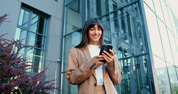 istock Young Caucasian businesswoman standing outside and smiling while using smartphone, with modern glass office building in the background 2244294174