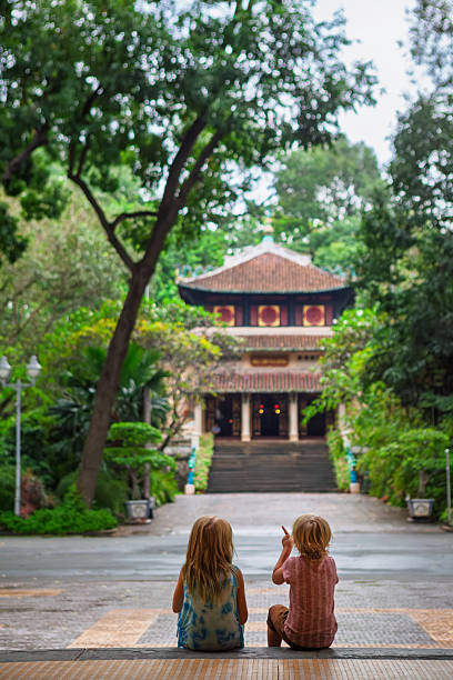 istock Two children sit on steps facing a traditional Vietnamese temple surrounded by lush tropical greenery in Saigon. Peaceful moment in cultural setting, perfect for family travel, and lifestyle themes. 2244305605