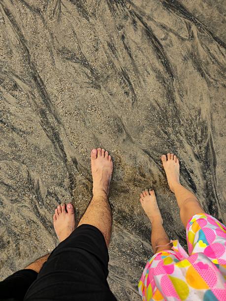 Closeup view of barefoot on wet sandy beach as gentle sea waves flow around feet. Soft sunlight highlights rippled sand texture and calm shoreline mood, capturing peaceful seaside moment of relaxation and connection.