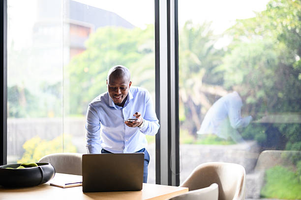 Cheerful professional man standing at a table while using a laptop and smartphone simultaneously in a modern office, with a backdrop of a large window and greenery.