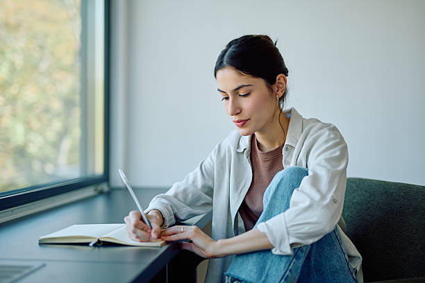 istock Young woman writing in notebook near window 2244326950