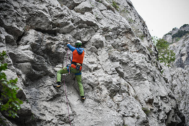 A lone climber wearing a green helmet, orange vest, and green pants climbs a rough limestone cliff skyward. A rope and safety gear secure the ascent, conveying focus, danger, and outdoor adventure.