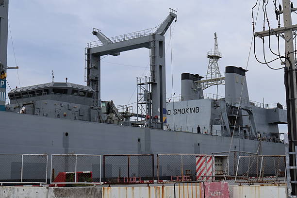 istock A large military ship docked at a harbor. The ship is quite sizable, with multiple decks visible above the waterline. It's painted in camouflage colors, typical for military vessels 2244341985