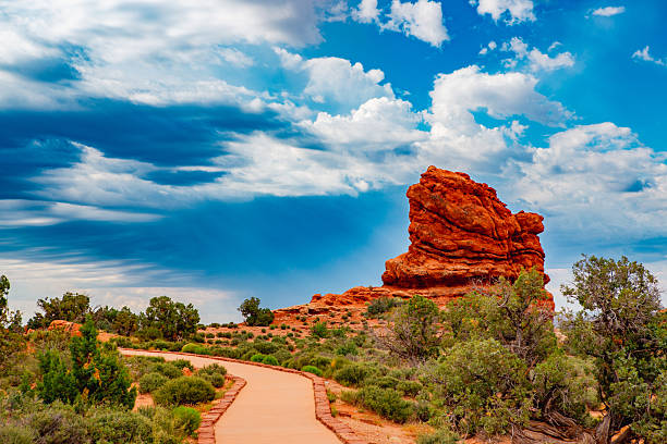 A solitary red sandstone formation towers against a turbulent desert sky, its contours glowing like embered iron under the filtered light of an approaching storm. A winding desert path — warm, smooth, deliberate — leads the eye directly toward the stone sentinel, symbolizing guidance, resilience, and the inevitability of challenge. The composition merges calm anticipation with impending power. The land is grounded in stillness, but the sky moves with energy and intent — the convergence of endurance and divine force.