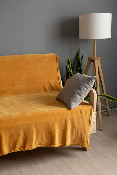 A mustard yellow sofa draped with a soft blanket sits in a modern living room, accompanied by a tall lamp and green plants for a stylish ambiance