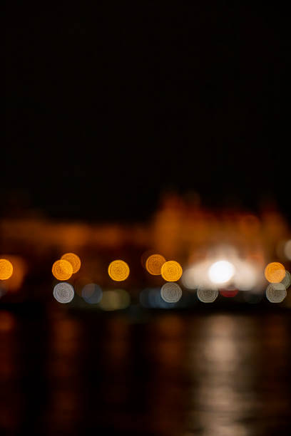 This image captures a blurred nighttime view of Hungarian Buildings in Budapest, Hungary, with its Gothic Revival architecture softly illuminated by warm orange lights. The lights create a bokeh effect, reflecting on the calm surface of the Danube River, evoking a serene urban nightscape. The dark water mirrors the glowing orbs, emphasizing the historic landmark against a black sky. No people are visible, focusing on the architectural glow and river tranquility. Ideal for commercial uses such as travel articles on Budapest tourism, illustrations of European city landmarks, backgrounds for Hungarian history content, or promotional materials for nighttime river cruises and urban exploration media.