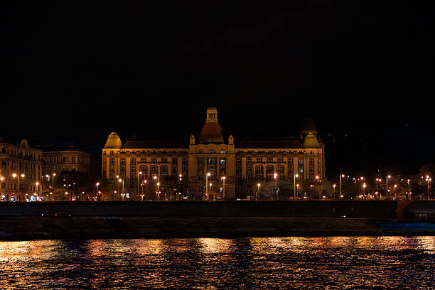 The iconic Hungarian Building stands prominently along the Danube River in Budapest, Hungary, captured in a nighttime scene with warm golden lights illuminating its Gothic Revival architecture featuring spires, arches, and ornate facades. The dark night sky contrasts with the building's glow, reflected on the calm river waters where a passenger boat glides by under nearby bridges. No people are visible in this serene urban landscape. This image suits commercial uses such as travel articles on European landmarks, tourism promotions for Budapest, historical architecture features, or illustrations in books about Hungarian heritage and nighttime cityscapes.