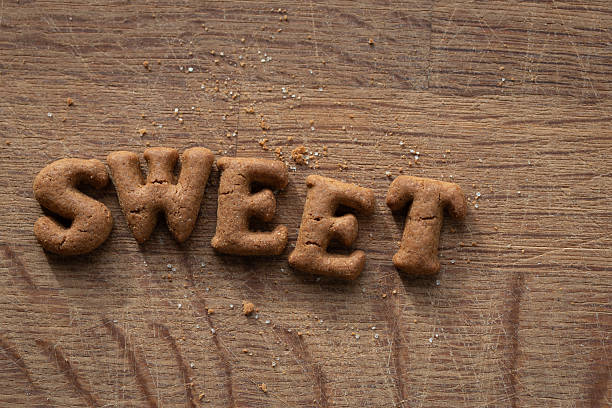 Close-up of the word “SWEET” created with small spiced cookies on a textured wooden table, with crumbs scattered naturally.