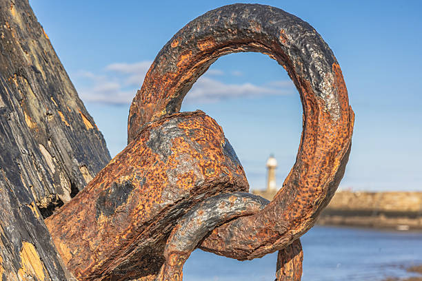 A close-up of an old rusting and disintegrating anchor ring and stock.  A lighthouse at the end of a pier can be seen in the distance.