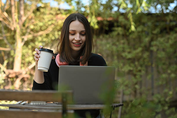 istock Young Caucasian woman enjoying remote work outdoors, smiling while using a laptop and holding a takeaway coffee cup 2244357049
