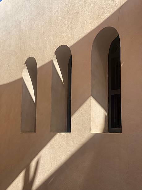 Soft natural sunlight casts geometric shadows across a beige stucco wall with three elegant arched windows, reflecting the simplicity and harmony of Middle Eastern architecture. The interplay of light and shadow creates a peaceful, minimalist aesthetic that captures the timeless beauty of desert design. Perfect for architecture, travel, interior design, and art projects emphasizing geometry, calmness, and modern heritage.