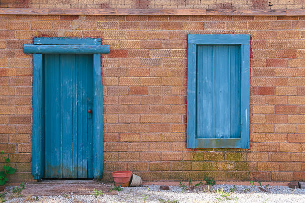 istock A weathered brick wall features a closed blue wooden door and a matching blue window shutter with flowerpots on the ground and scattered green plants in front 2244365372
